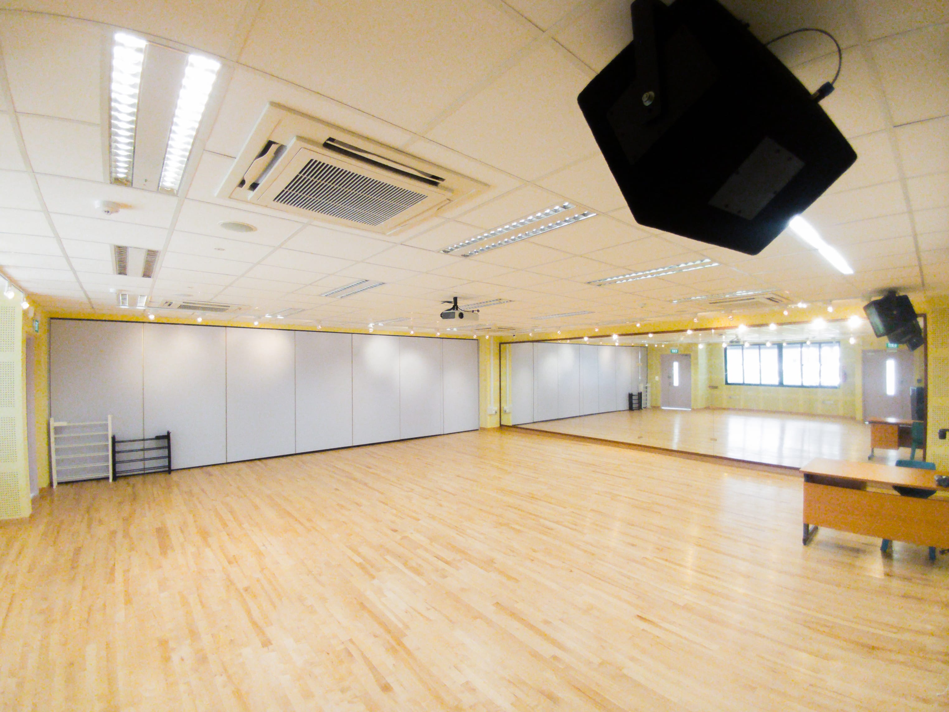 Empty dance studio with light wood floors, white partition walls, ceiling projector, speakers, and a large mirror on one wall.